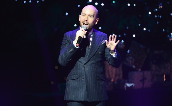 A bald man in formal attire singing into a microphone, engaging the audience with his performance.