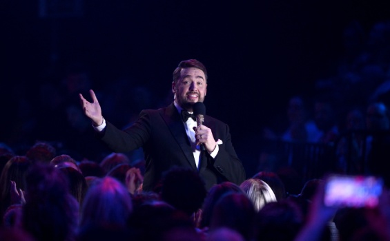 A man in a tuxedo stands confidently, holding a microphone, ready to speak or perform at an event.